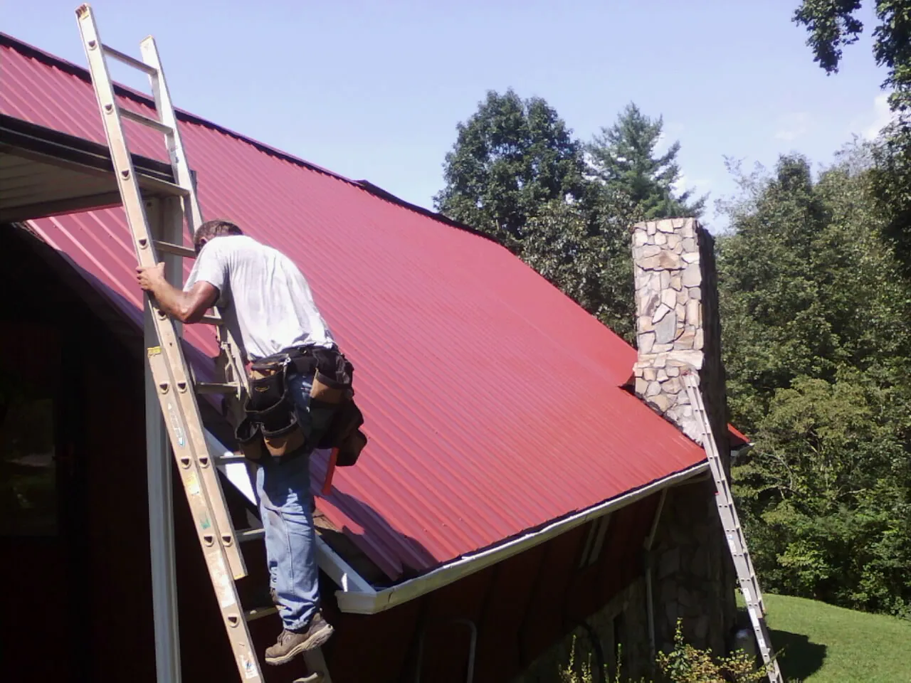 Roofer on a ladder beside a red metal roof and stone chimney — Soffit & Fascia Repair by Roofing Brothers in Spencertown